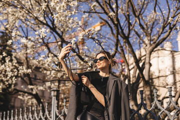 Fototapeta na wymiar Focus on phone. Happy young woman photographing herself using her mobile phone. Caucasian female talking selfie with her smart phone on blooming tree background.