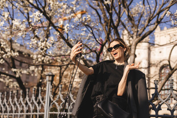 Fototapeta na wymiar Happy young woman photographing herself using her mobile phone. Caucasian female talking selfie with her smart phone on blooming tree background. Girl posing in sunglasses, suit, t-shirt, pants, bag.