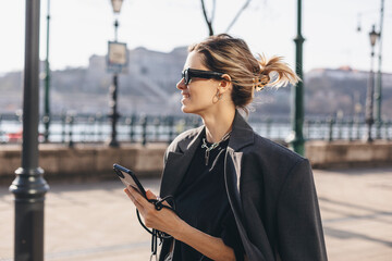 Beautiful young blonde girl talking with friends on a video call walking in the city. Smiling woman...