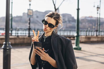 Happy young woman photographing herself using her mobile phone. Caucasian female taking selfie with her smart phone at city. Woman show peace sign and blow kiss, talk by video call, wear grey jacket.