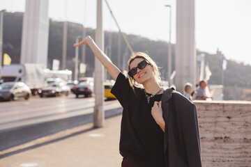 Pretty curly woman in sunglasses enjoying city view from bridge in sunny day. Lovely blonde woman...