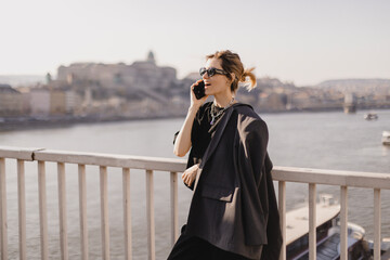 Woman talking on the phone walking on the bridge under the river. Beautiful young woman with blond hair smiling and having a conversation over the phone in the city. Business woman on smartphone.