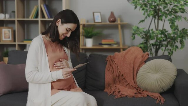Caucasian Woman in Expectation Holding Ultrasound Scan and Touching Belly While Sitting on Comfy Sofa. Young Lady Enjoying Future Motherhood with First Sonogram Image of Unborn Baby at Home