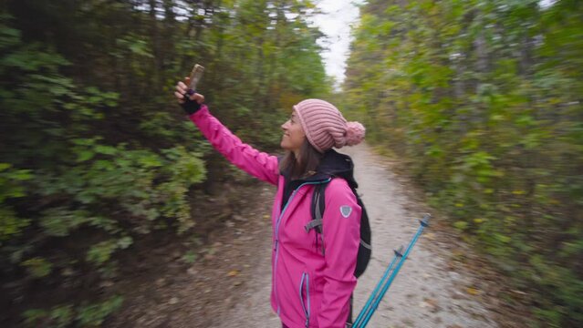 Hiking girl with poles and backpack on a dirtroad or trail in mountain forest. Taking selfy photo with her cell phone. Travel and healthy lifestyle outdoors in fall season. Circular camera movement..