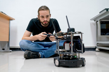 Smart Caucasian professional engineer or technician worker man sit on floor use controller to move small robotic machine that put on the floor in room of factory workplace.