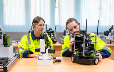 Two Caucasian professional technician or engineer workers sit and help to check and maintenance small robotic machine on table in factory room workplace.