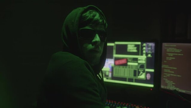 Black Hooded and Glasses-Wearing Mysterious Hacker or Programmer Hacking in an Abandoned Room with Green Lights Suddenly Turns to Look at the Camera, Close-Up Shot