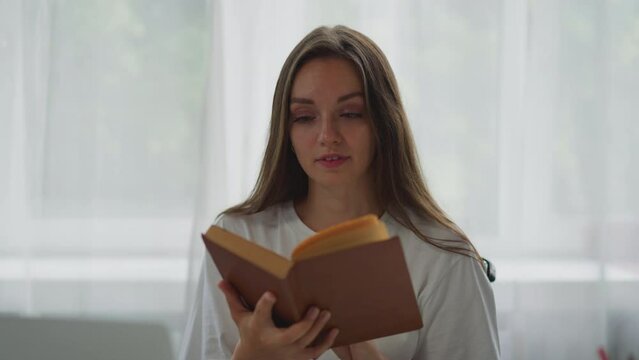 Focused woman sits in wheelchair reading interesting book against bright window with white curtain. Lady smiles enjoying story closeup