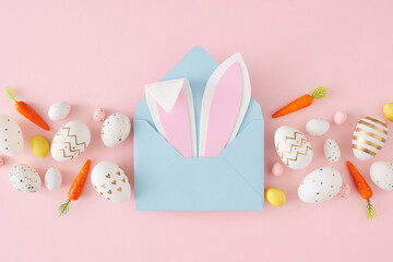 Easter decor concept. Top view photo of open blue envelope with easter bunny ears colorful eggs and carrots on isolated pastel pink background