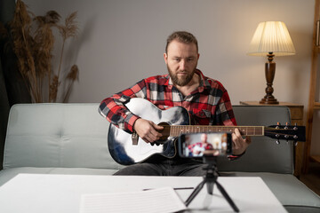 A man with acoustic guitar plays music live staying home in front of the smartphone camera.