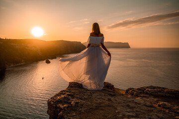 Sunset woman dress. A happy woman in a white light dress stands on the edge of a cliff above the...