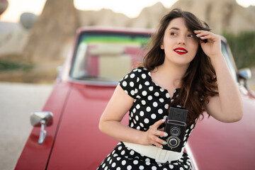 beautiful girl in retro style posing near vintage red cabriolet car with old film camera in hands.