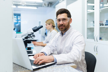 Naklejka na ściany i meble Portrait of mature successful scientist lab technician at workplace in laboratory with microscope, senior man with beard and goggles smiling and looking into camera, inventor with laptop .