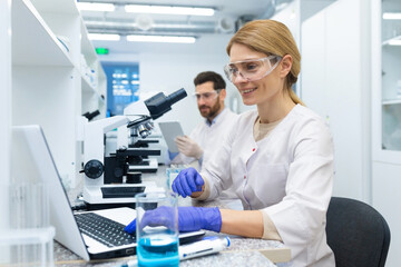 Mature female scientist in white coat at work, laboratory assistant using microscope and laptop at work, scientist working inside laboratory researching.