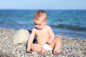 Fototapeta na wymiar Portrait of an infant in a diaper sitting on a pebble beach against the background of the sea and played with pebbles