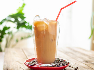 a drink sitting on top of a red plate with coffee beans on top of a wooden table, plant on background. Iced latte in summer cafe