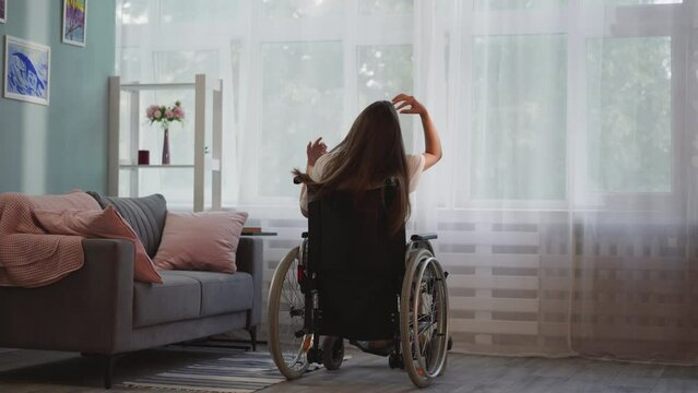 Lonely lady with disability with multiple sclerosis looks out bright window in room. Brunette woman sits in wheelchair touching long loose hair backside view