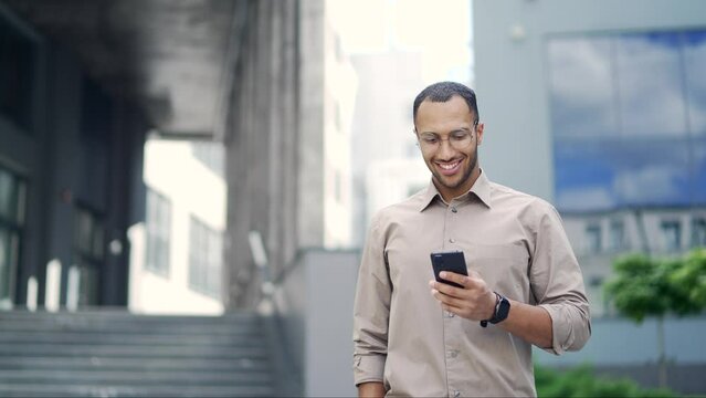 A smiling young adult man is using a smartphone while walking on the street near an office building. Happy handsome mixed race male checking email, chatting online, browsing social media, texting