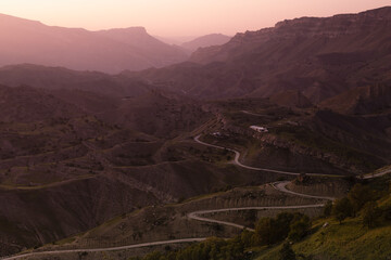 Idyllic landscape  - silhouette of high mountain green ridges in pink pastel sunrise sunlight, mist, serpentine road, village away in early summer morning, panorama view. Majestic travel in Dagestan.