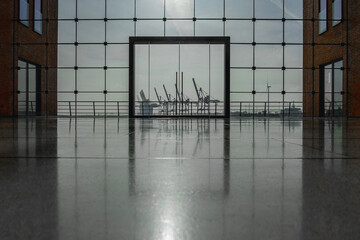 Blick durch ein großes modernes Glasfenster, mittig eine große Glastür, links und rechts rotbraune Gebäudeteile, im Hintergrund die Silhouetten von Hafenkränen und Industriegebäuden zu sehen, horizont