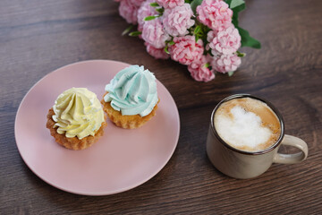 Obraz na płótnie Canvas Two delicious, Sweet cream Cakes on a pink plate with yellow and blue cream sit on a wooden table. A cup of cappuccino with wooden Capcakes on the background. Close-up. Copy space