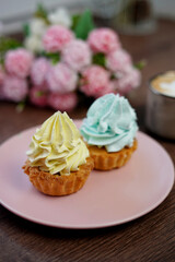 A Pink Plate with birthday Capcakes sits on a wooden table. A beige cup of cappuccino sits on the table. Near a plate of cakes lie beautiful pink and white flowers.