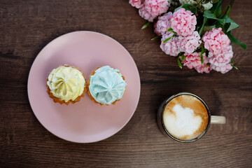 A top view of Two yellow and blue Cream Capcakes on a pink plate sit on a Wooden Table. A beige cup...