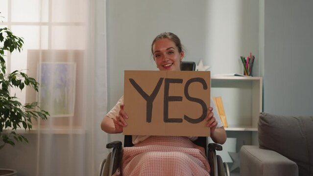 Woman with paralysis shows poster with inscription Yes smiling and looking in camera. Young female person with disability sits in wheelchair after accident