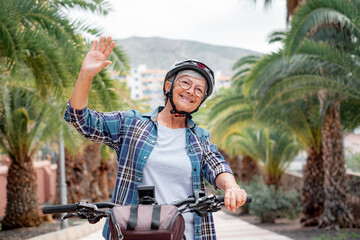 Handsome senior woman with helmet riding her electric bicycle in park waving hand. Elderly smiling...