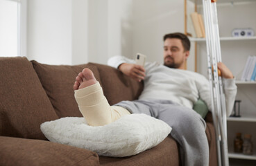 Man with a broken leg using his mobile phone while relaxing on the sofa at home, with his foot...
