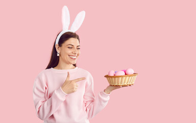 Happy beautiful woman wearing pink sweatshirt, earrings and bunny ears standing on pastel pink studio background, smiling and pointing at basket of pink Easter eggs that she is holding in her hand - Powered by Adobe