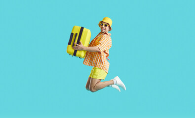 Crazy happy young man going on summer vacation high jumps with suitcase on light blue background. Excited guy in summer shorts, shirt, panama and sunglasses having fun with suitcase in his hands.