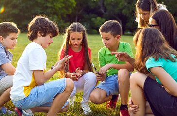 Kids learn about environment during field trip. Students have interesting biology class in nature. School children together with teacher sit down on green grass, search for insects, look through loupe