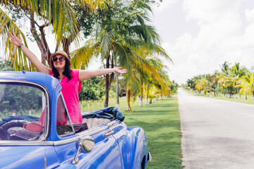 Happy female traveler in sunglasses and hat standing in blue vintage cabriolet with raised arms...