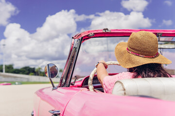 Anonymous female traveler in straw hat driving pink retro cabriolet on cloudy day during summer...