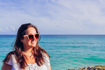 Happy female tourist in sunglasses and white summer dress smiling brightly while looking away on sandy beach against blue sea during vacation in Cuba. Cheerful woman in sunglasses on beach