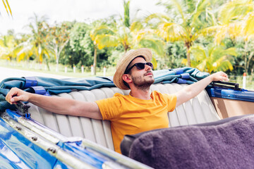 Relaxed male traveler in sunglasses and hat sitting in cabriolet with arms outstretched while...