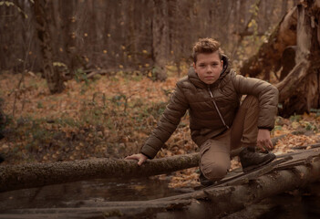 Boy sits on the wooden bridge over the river in fall forest. Brown toned.