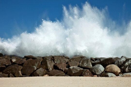 Ola rompiendo con furia en un dique de rocas con cielo azul.