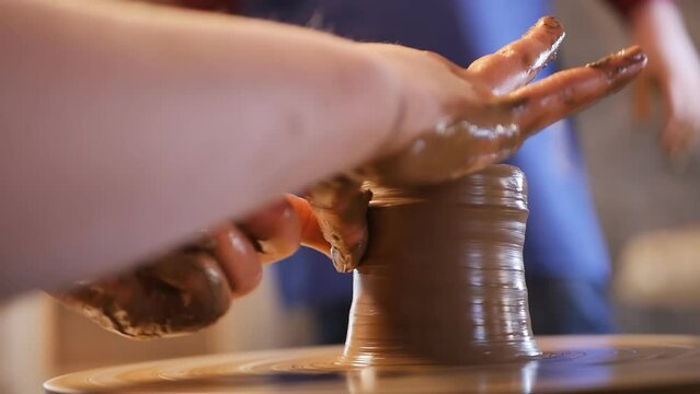 Pottery lesson. The hands of a teenage student are making a clay vase on a rotating potter's wheel and the hands of an adult male teacher are helping to make a pasuda of clay while teaching a child