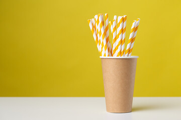 Disposable cardboard biodegradable cup on a white table with a straw against a yellow background.
