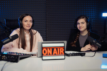 Female host and guest sitting at table with equipment and tablet with On Air inscription while...
