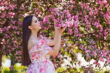 Obraz na płótnie Canvas A beautiful girl with long hair, standing near pink blooming apple trees, holding a blooming branch