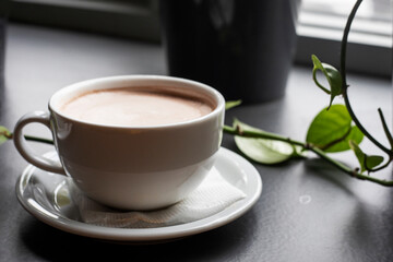 white cup with hot coffee latte on the windowsill with green ivy. horizontal