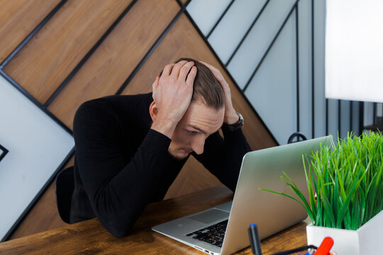 A businessman is sitting at his desk, looking at his laptop and holding his head due to some problems that have happened.
