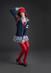Young woman in preppy outfit and red cloche hat holding down short skirt isolated on gray background - 575449093