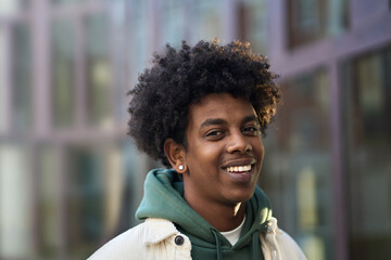 Smiling positive young African American guy model standing at big city street. Stylish ethnic hipster gen z teenager boy feeling happy looking at camera outdoors, close up portrait. - obrazy, fototapety, plakaty