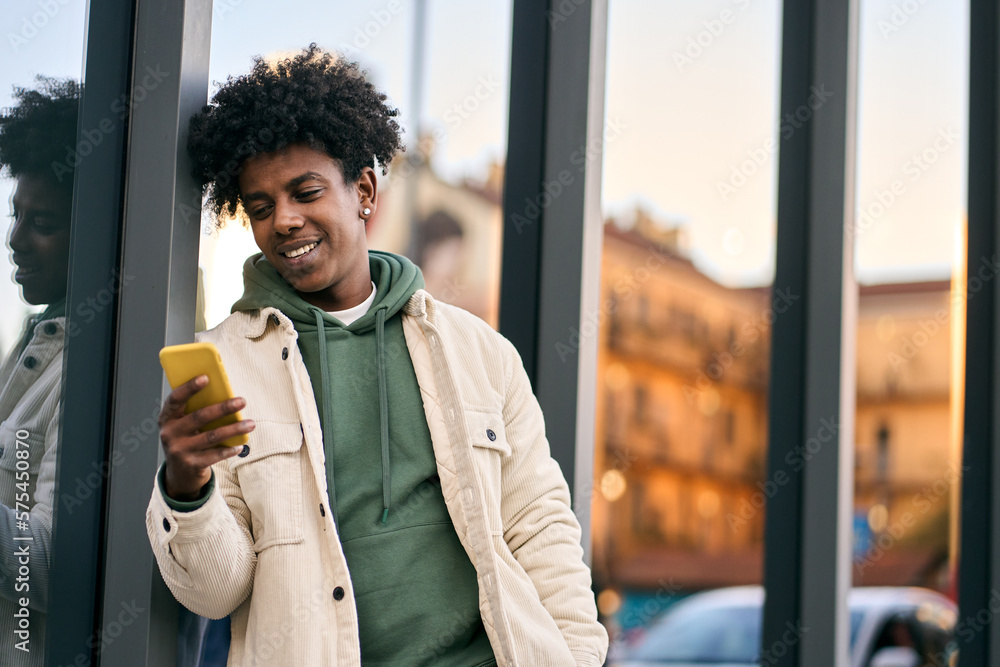 Wall mural cool smiling young african american teen guy holding mobile phone standing at glass city wall. happy