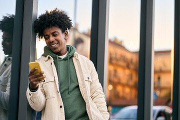 Cool smiling young African American teen guy holding mobile phone standing at glass city wall....