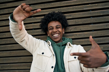 Young happy African American hipster teen guy laughing pretending taking selfie at wooden wall...